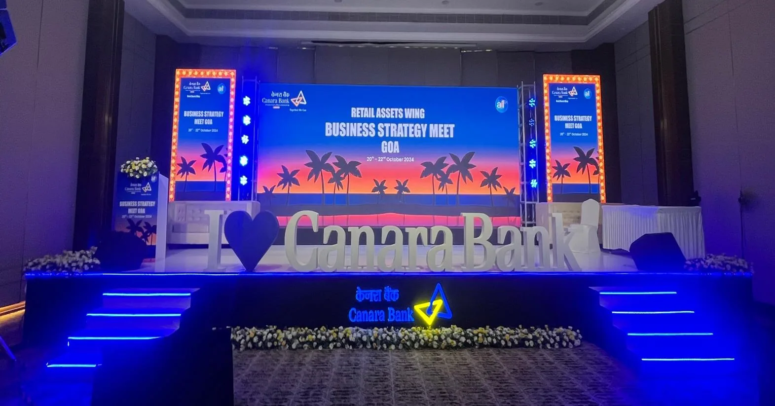 Main conference stage for the Canara Bank Business Strategy Meet - wide LED backdrop with a palm-and-sunset beach graphic reading 'RETAIL ASSETS WING BUSINESS STRATEGY MEET GOA', flanked by bulb-lit vertical banner towers, oversized white 3D 'I love Canara Bank' letters on the riser, blue LED-lit steps and a flower-bed apron