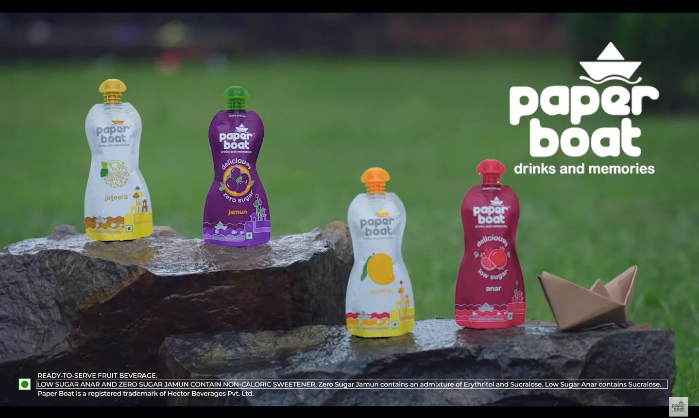 Campaign film end frame: four Paper Boat pouch packs - jaljeera, zero sugar jamun in purple, aamras in white and orange, and low sugar anar in magenta - arranged on dark wet rock ledges against a soft-focus green lawn, with a folded kraft paper boat at right and the large white Paper Boat 'drinks and memories' logo reversed over the grass; a statutory ready-to-serve beverage and sweetener disclaimer strip runs across the black lower band